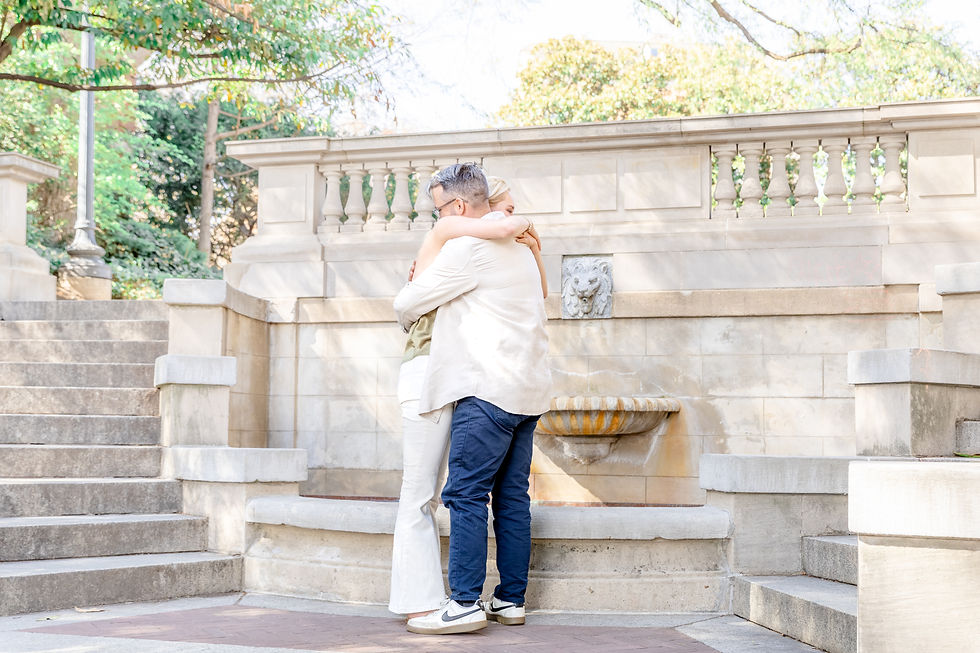 A couple hugs tenderly by a stone fountain in a sunlit park. Stone steps and greenery surround them, creating a serene, intimate scene.