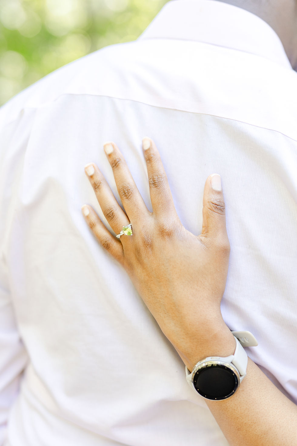 A hand with a ring gently rests on a person's white shirt back. The hand wears a white smartwatch. The background is softly blurred green.