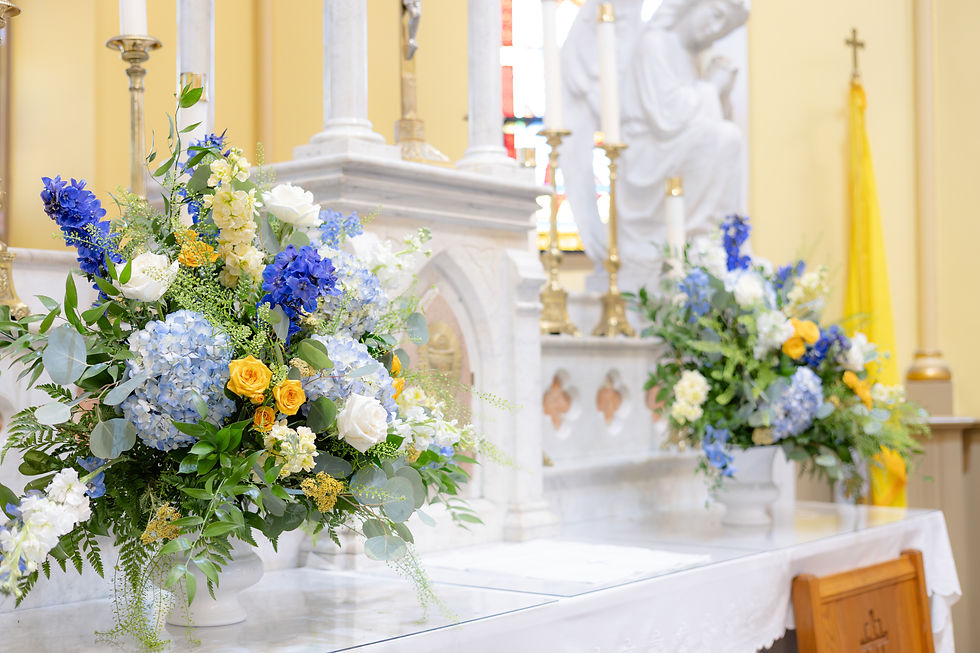 Flower arrangements with blue, yellow, and white blooms on a marble altar in a church, featuring angel statues and stained-glass windows.