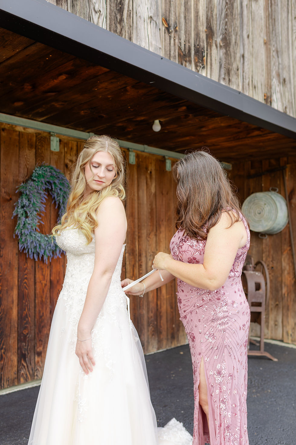Woman in pink sequined gown laces a bride into her white wedding dress beside a rustic wooden wall.