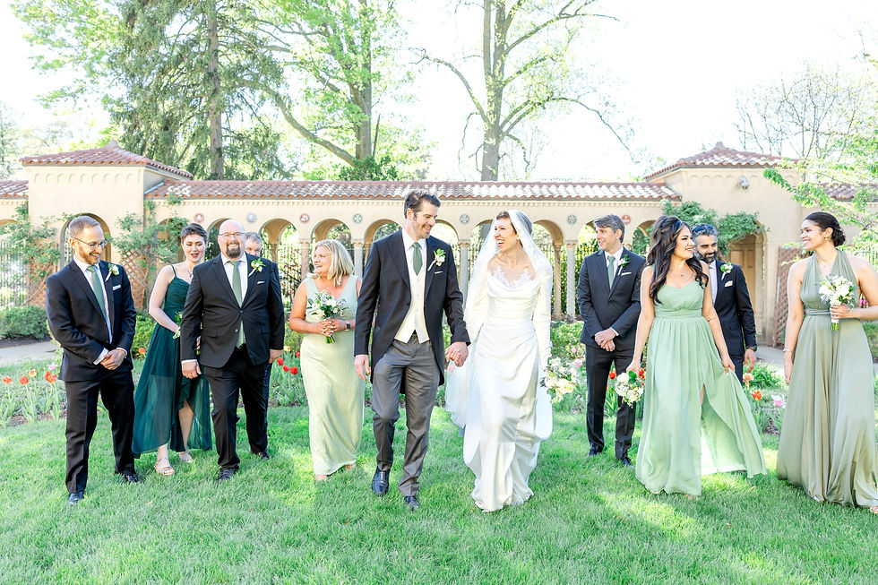 Bride and groom walk joyfully with wedding party in elegant attire. Green garden setting with blooming flowers and bright sunlight.