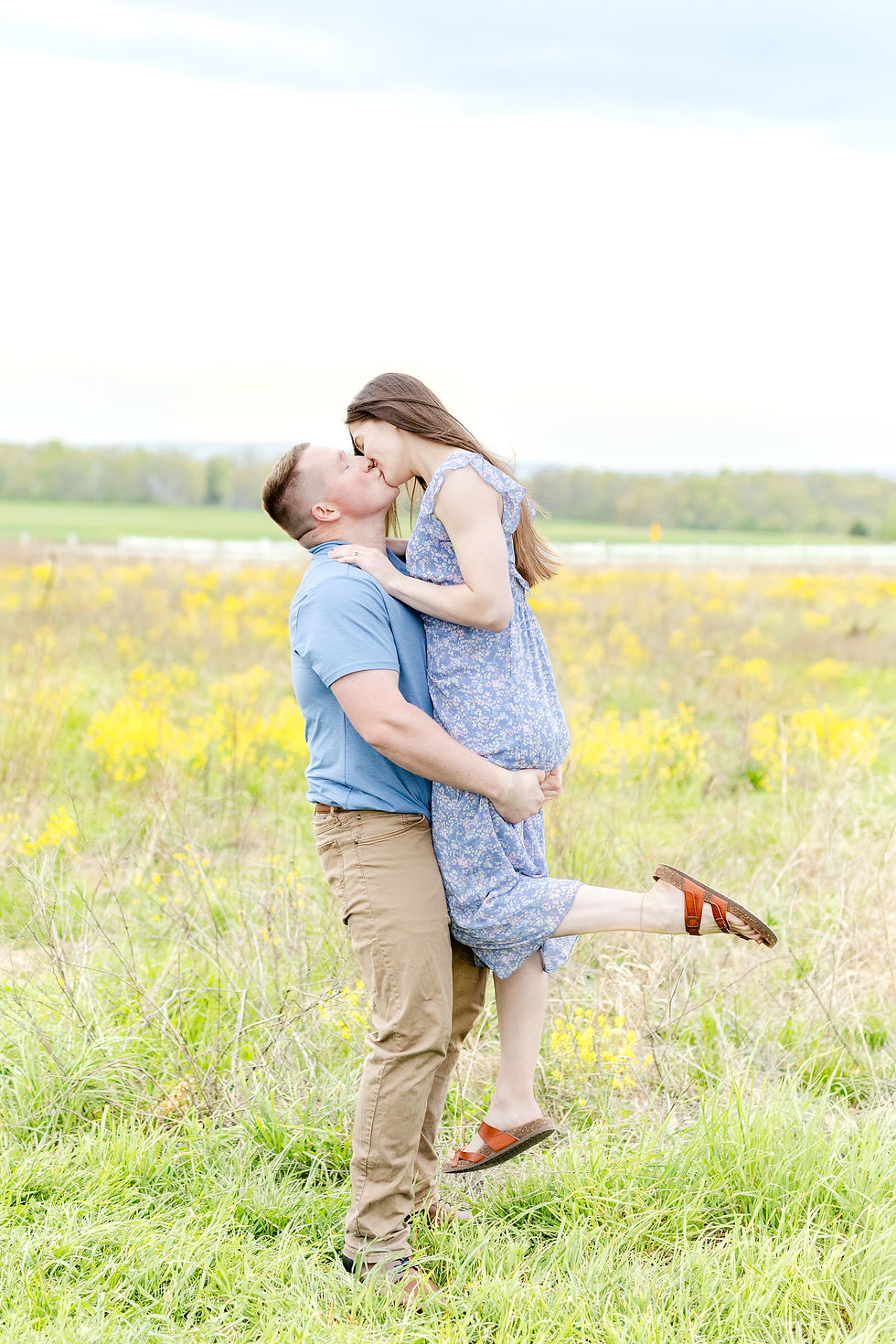 A couple kisses in a sunny field. The woman in a floral dress is lifted by the man in a blue shirt, surrounded by green grass and yellow flowers.