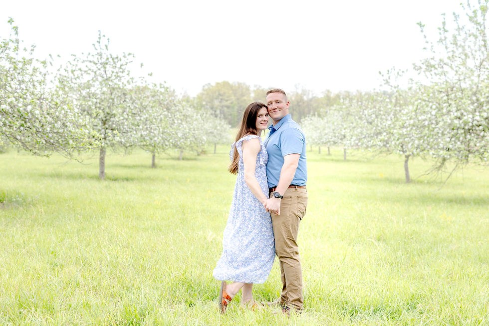 A couple holding hands, smiling in a blooming orchard. Woman in a blue floral dress, man in a blue shirt with tan pants. Bright, airy mood.