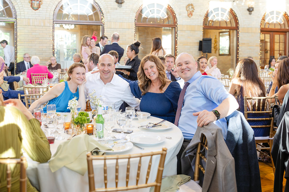 Smiling group of four pose at a round table with white tablecloths in a festive hall. Cheerful atmosphere, others mingling in the background.