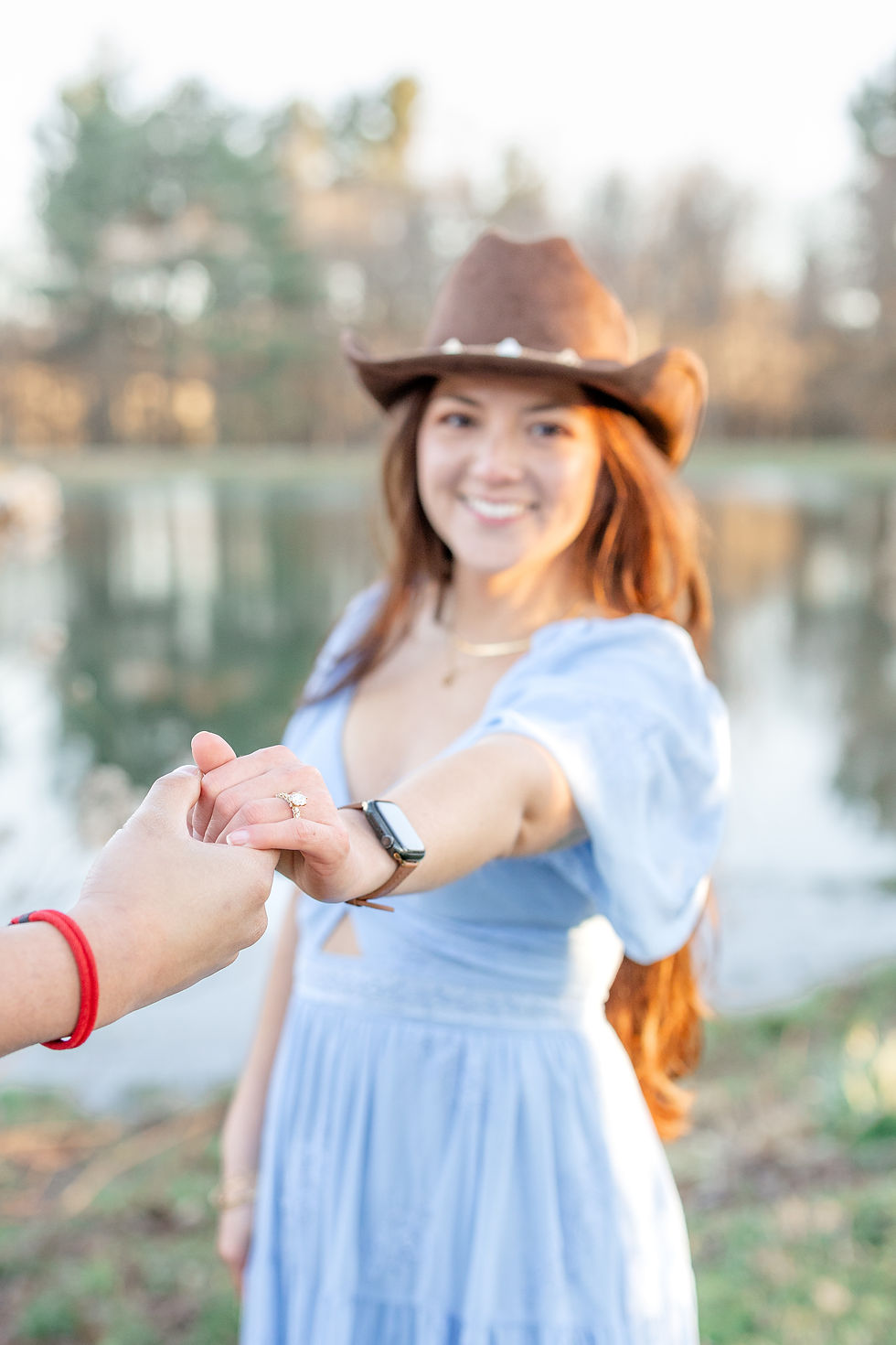 Woman in a cowboy hat and blue dress holds hands, showing an engagement ring. Smiling near a pond with trees in the background.