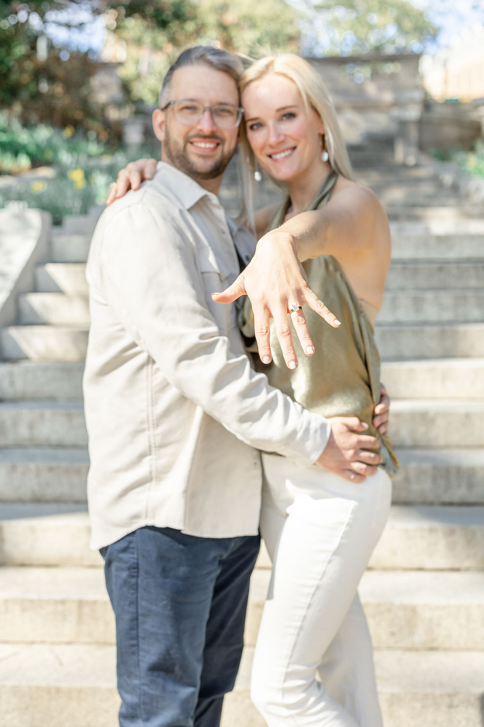 Couple smiling on stone steps, woman showing engagement ring. She's wearing a green top and white pants; he's in a beige shirt and navy pants.