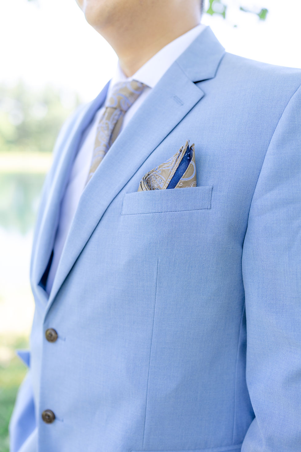Close-up of a man in a light blue suit with patterned tie and pocket square, standing outdoors in bright daylight.