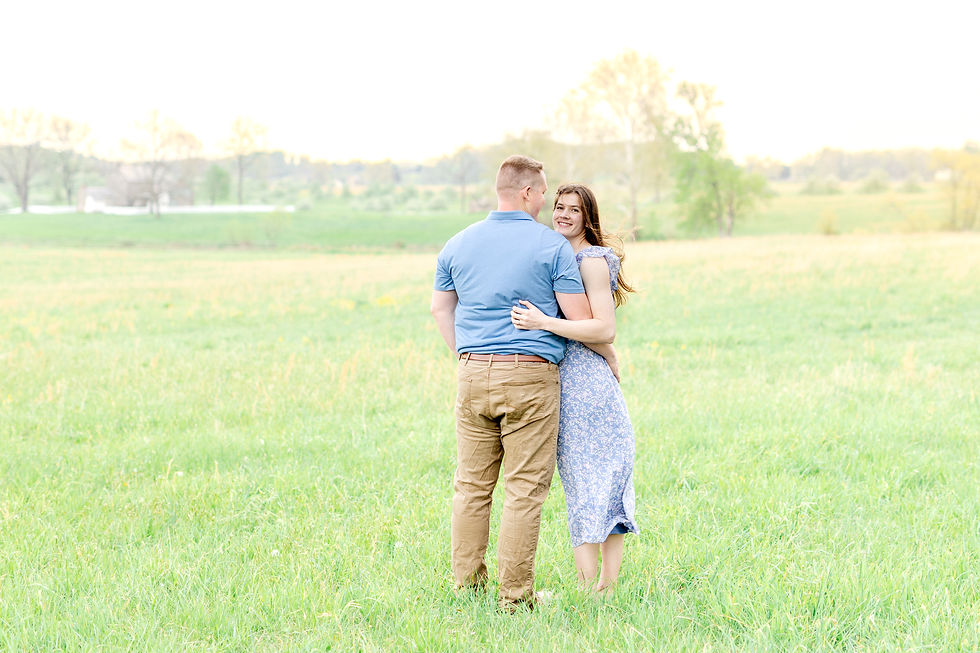 A couple embraces in a sunny field. The woman smiles, wearing a blue floral dress; the man in a blue shirt and tan pants. Lush green grass.