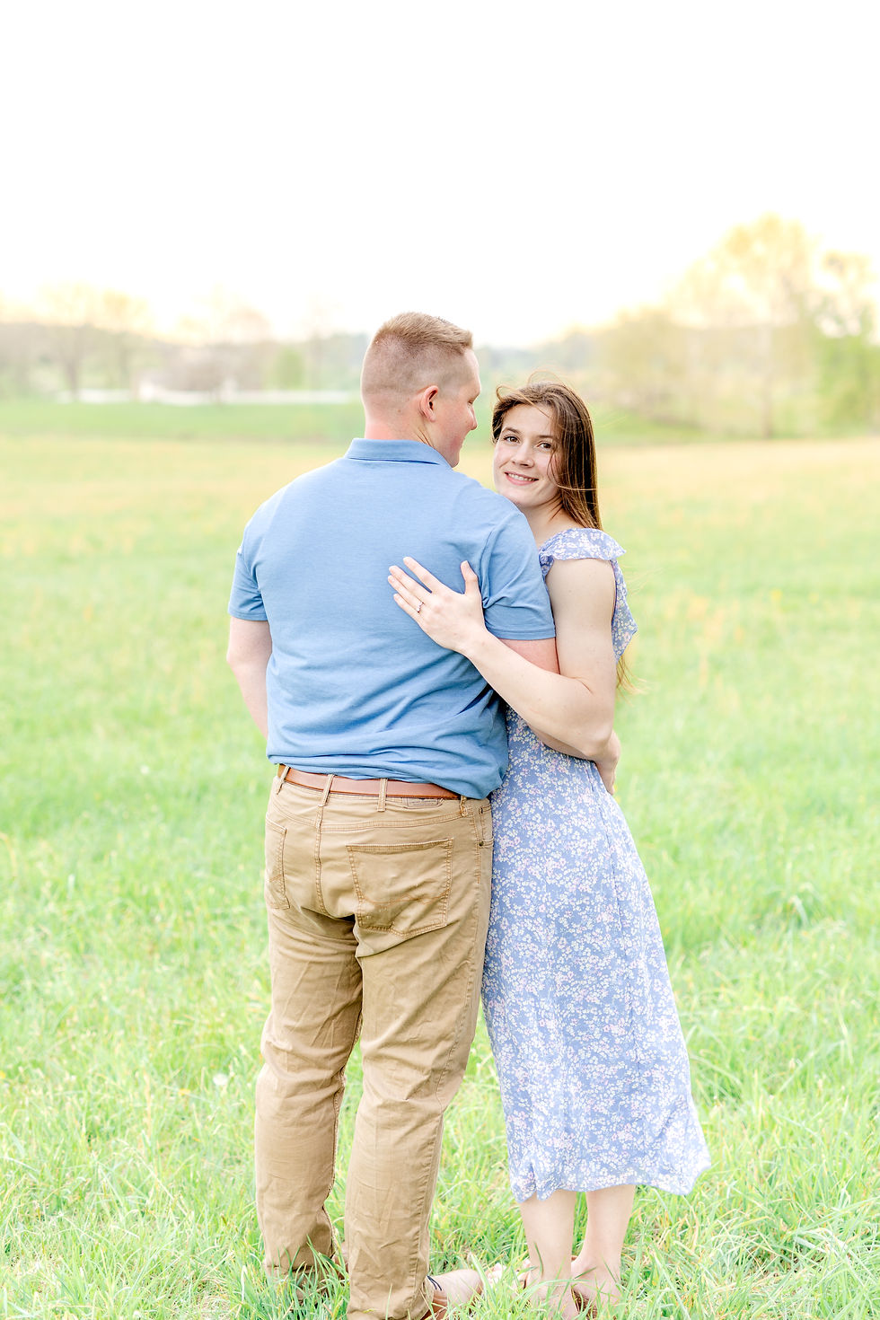 Couple embraces in a sunny field. Woman in blue floral dress smiles at camera. Man in blue shirt and khaki pants. Serene, joyful mood.