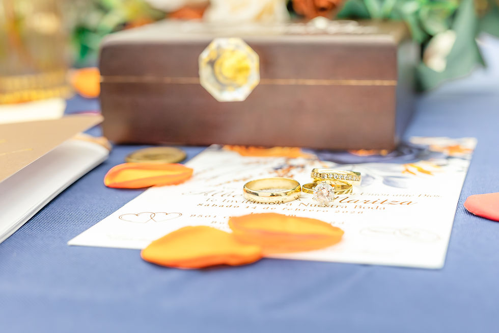 Wedding invitation with two gold rings on a blue tablecloth. Orange petals scattered around, with a wooden box in the background.