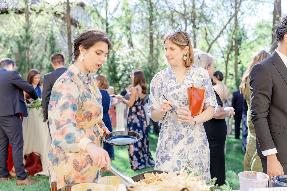 Women in floral dresses at an outdoor buffet; one serves herself while chatting with another. Guests mingle in a sunny garden setting.