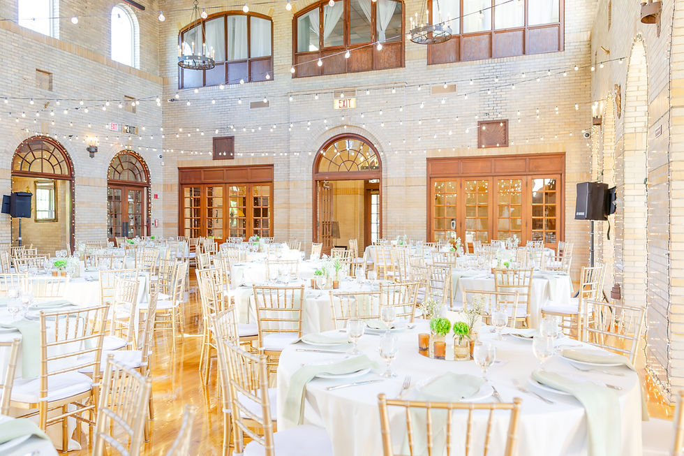 Elegant event hall with round tables, white tablecloths, gold chairs, and green accents. String lights create a festive atmosphere.