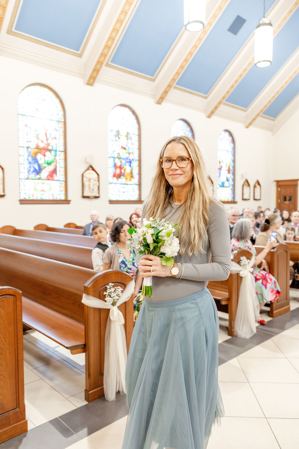 Woman in a grey top and blue skirt holds white flowers, walking down a church aisle with wooden pews and stained glass windows.