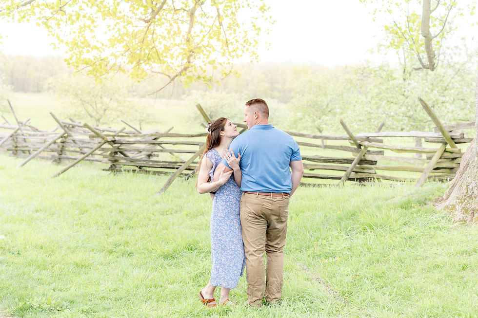 A couple embraces in a grassy field by a wooden fence. The woman wears a blue dress, and the man wears a blue shirt. Trees and soft light surround them.