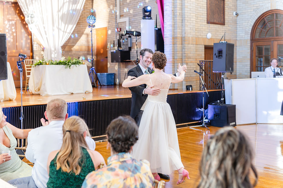 A couple dances joyfully at a wedding reception with guests watching. Brightly lit hall, wooden floors, and elegant decor in the background.