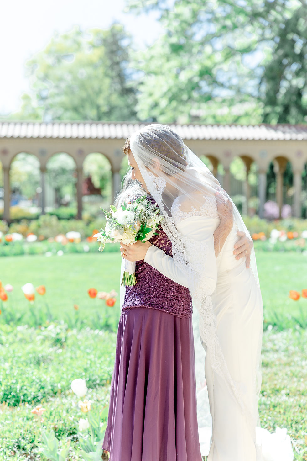 Bride in white gown and veil hugs woman in purple dress, holding bouquet. Bright garden with arches and flowers in background.