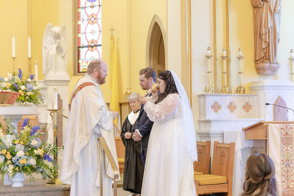 Bride receiving communion from a priest in a church with stained glass. Groom and a nun stand nearby. Floral arrangements add color.