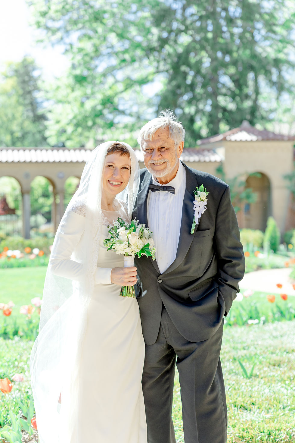 Bride and groom smiling in a garden setting. Bride holds a bouquet of white flowers; groom wears a black suit with a boutonniere. Sunny day.