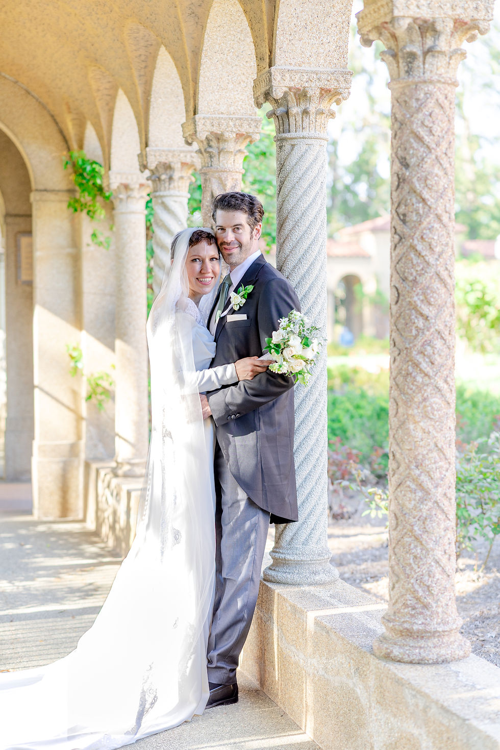 Bride and groom embrace under ornate archway. Bride in white dress, groom in black suit, both smiling. Sunlit garden background.