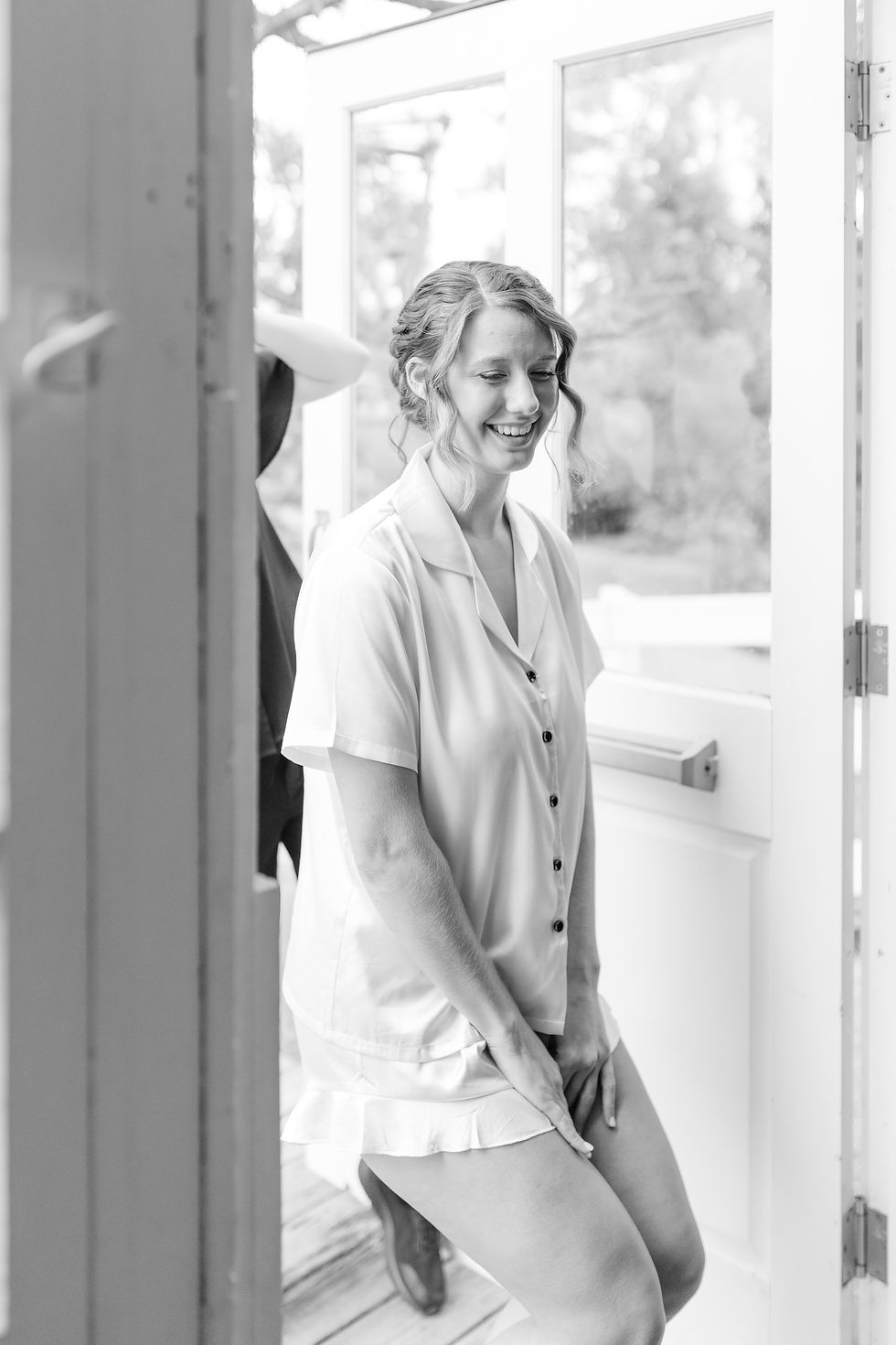Black-and-white photo of a smiling woman in light pajamas standing in a doorway, with another person partly visible behind her.