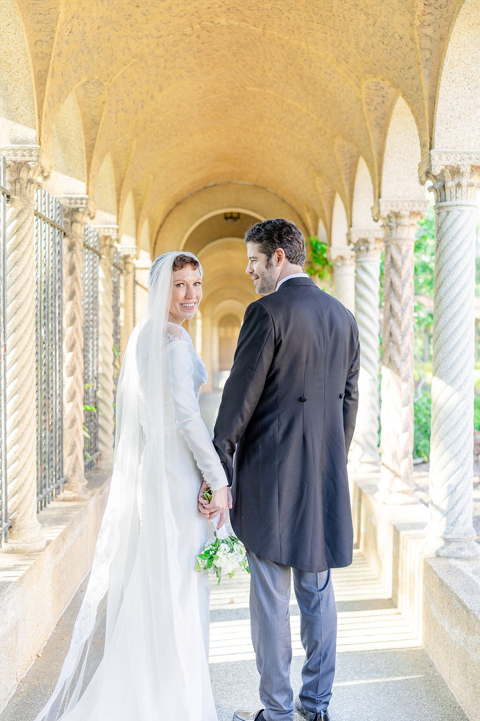 Bride and groom holding hands, smiling in an elegant stone archway. Bride in white gown, groom in black suit. Sunlit, romantic setting.