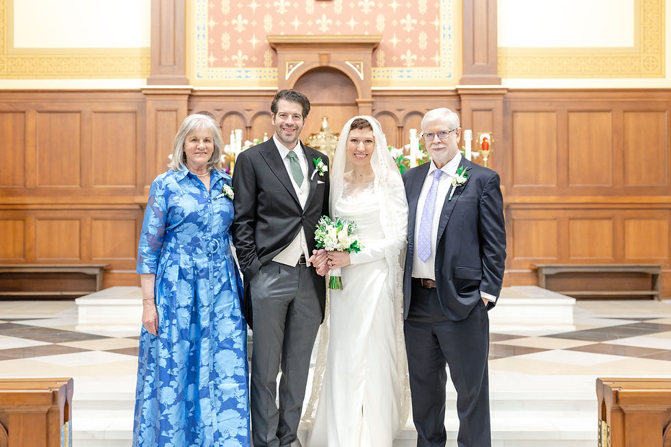 Bride and groom with parents in a church, smiling. Bride in white dress, groom in suit, mom in blue dress, dad in suit. Ornate wood altar.