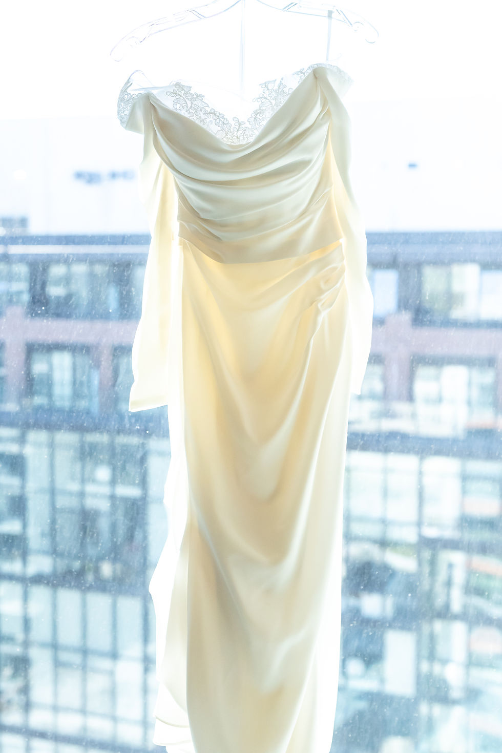 Elegant ivory wedding dress with lace trim hangs on a hanger against a window, city buildings visible in the soft, bright background.