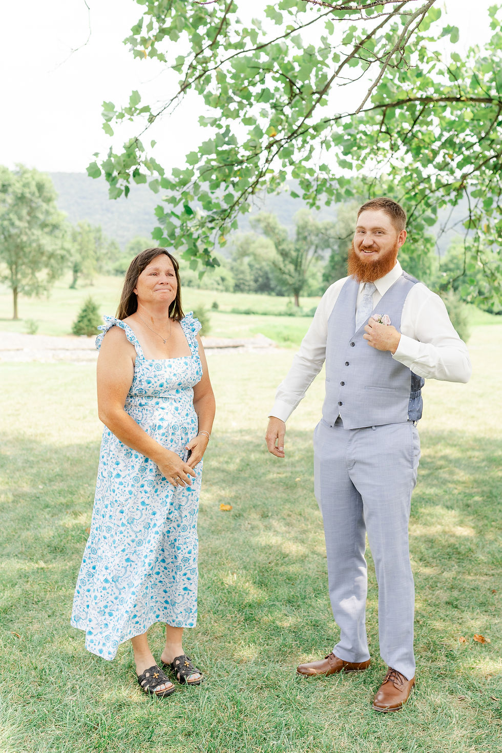 Woman in a blue floral dress and man in a light gray suit stand smiling under a tree in a sunny park.