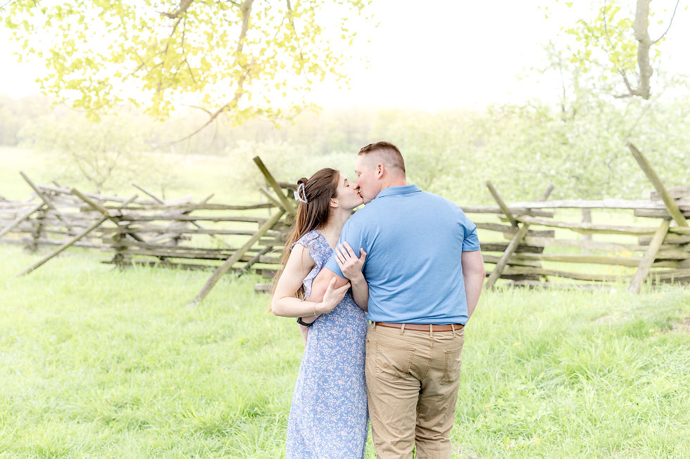 A couple kisses in a sunny field by a wooden fence. The woman wears a blue floral dress, the man a blue shirt. Peaceful, romantic setting.