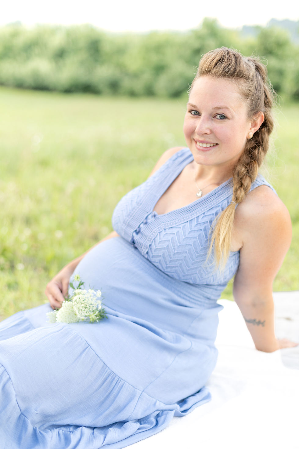 Pregnant woman in a blue dress sits on a blanket in a grassy field, smiling. She holds flowers and the mood is serene and joyful.