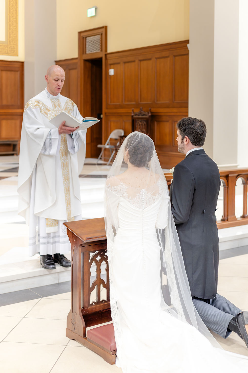 A couple kneels before a priest holding a book during a wedding ceremony. The bride wears a white dress with a veil in a church setting.