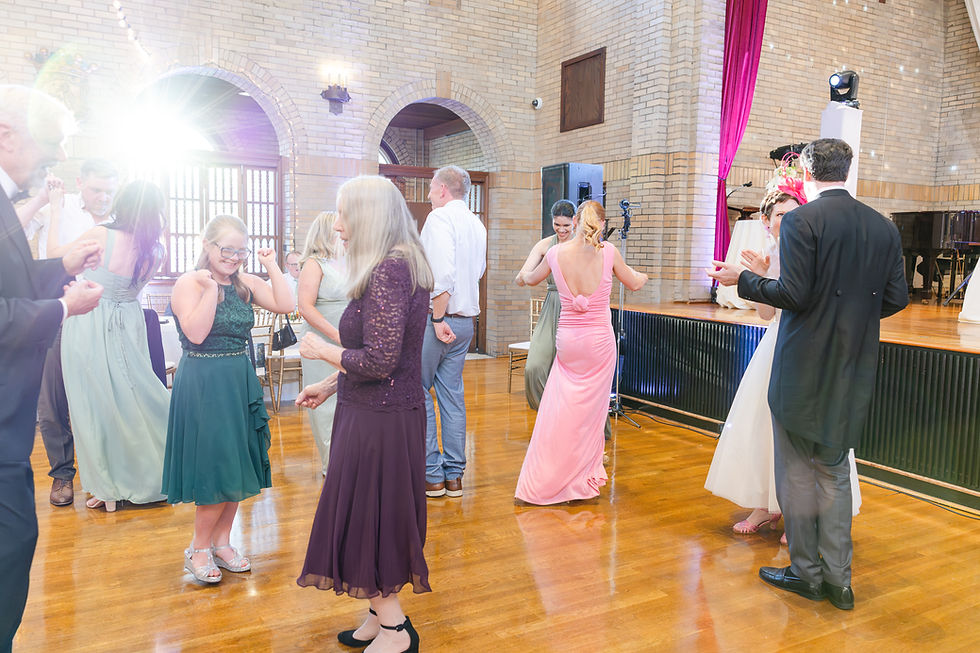People dancing in a bright hall with wooden floors and brick walls. Women in colorful dresses and men in suits. Festive atmosphere.