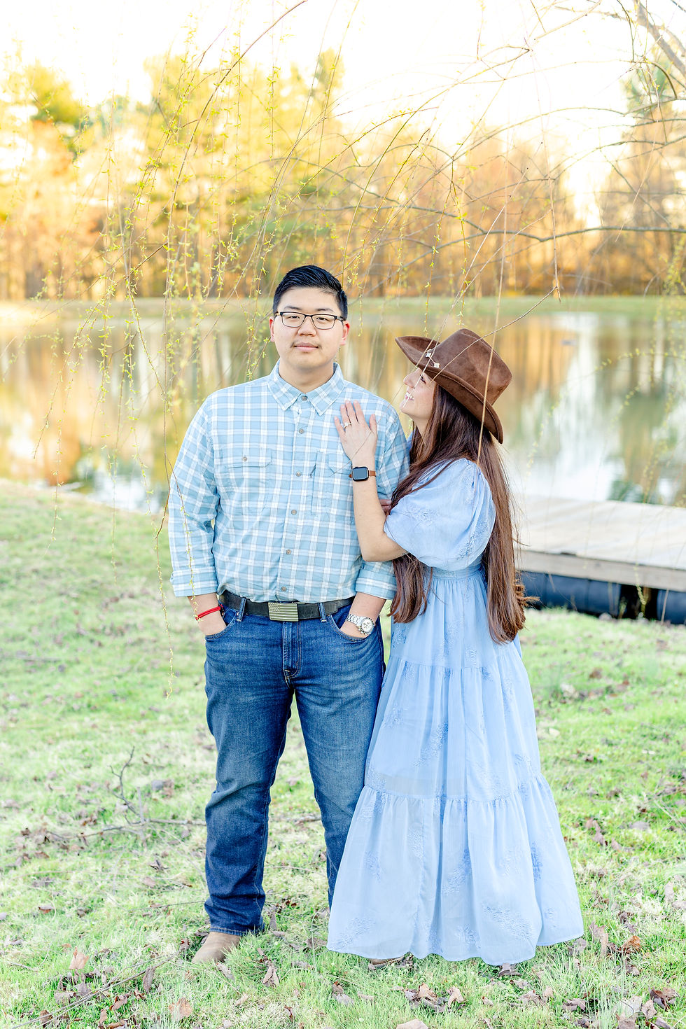 A couple by a pond; man in plaid shirt, hands in pockets. Woman in blue dress, brown hat, hand on his shoulder. Trees and water behind.