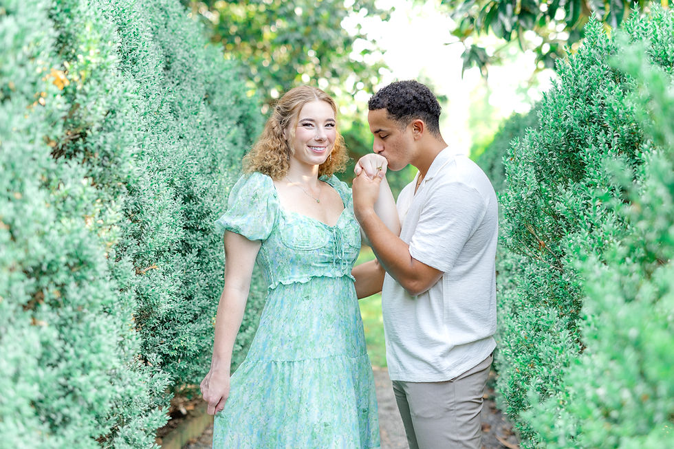 Smiling couple in a lush garden, man kissing the woman's hand while she wears a light green dress.