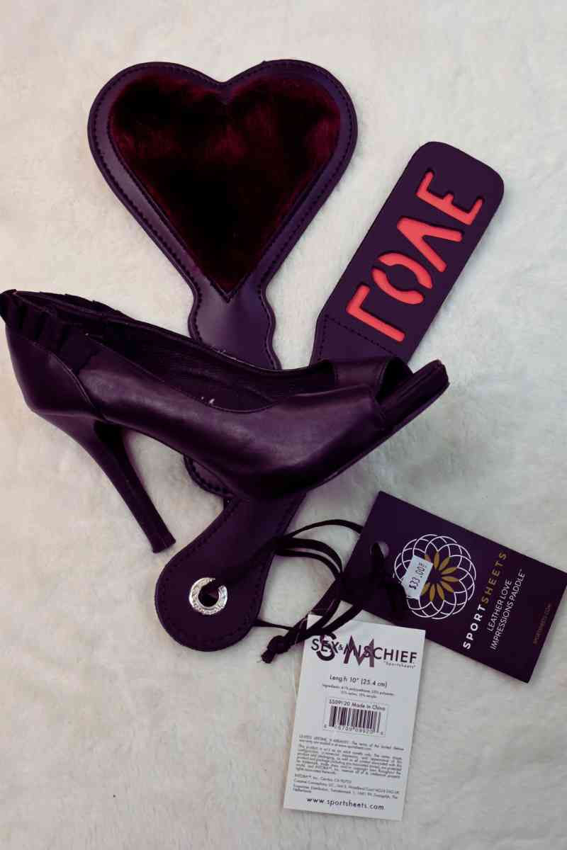 A collection of BDSM accessories including a heart-shaped paddle, a black stiletto shoe, and other fetish gear, perfect for impact play and available at a sex shop