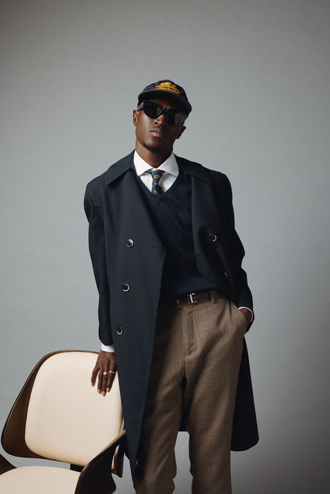 A stylish man leans casually against a modern chair, dressed in a sharp black trench coat over a v-neck sweater, tie, and tailored brown trousers. He accessorizes with a navy cap, dark sunglasses, and exudes a cool, contemporary vibe against a neutral background.