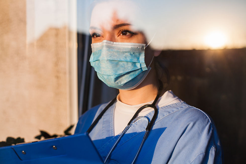A dedicated female physician in scrubs and a mask gazes thoughtfully through a window, highlighted by the warm glow of the setting sun.