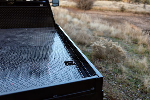 A black powder coated diamond plate flatbed with bed rails and inset D-rings parked in a field.