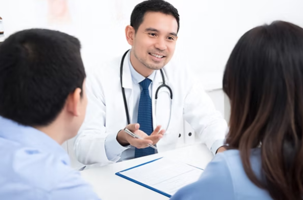 Smiling doctor in white coat with stethoscope talks to two patients across a desk in a bright clinic.