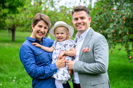 Familienportrait. Mutter und Vater halten ihre Tochter zwischen sich. Alle schauen lachend in die Kamera. Sie stehen auf einer Wiese mit Apfelbäumen.