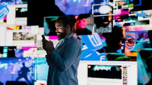 A bearded man in a blue shirt scrolls a smartphone, face lit by the screen glow, standing in front of a wall of overlapping social media videos and digital content.