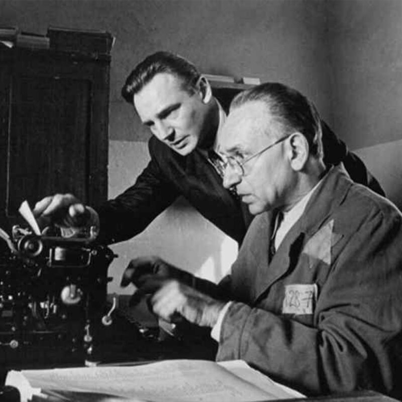 Dos hombres discuten sobre una máquina de escribir en una oficina en blanco y negro. Uno teclea mientras el otro observa con interés.