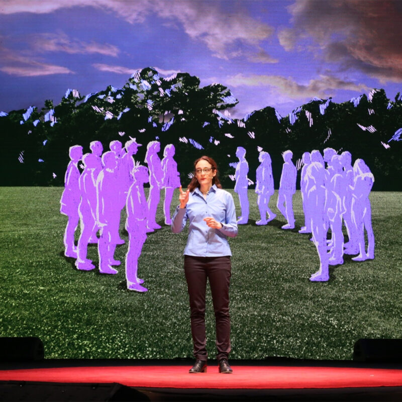 Una persona presenta en un escenario con fondo de siluetas púrpuras y naturaleza. Viste camisa azul y parece estar hablando.