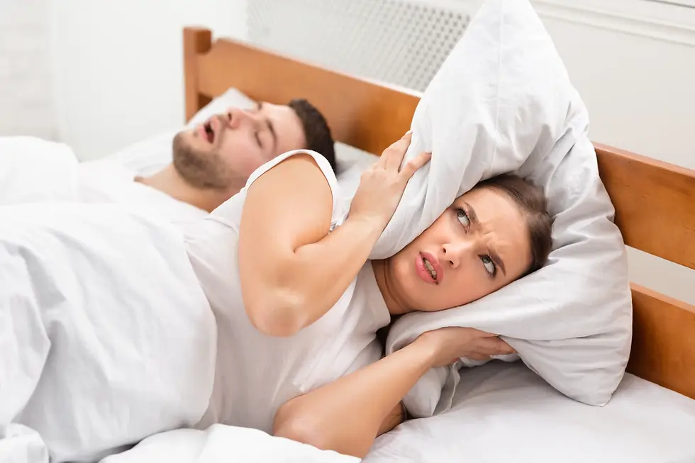 A couple in white pajamas lies on a bed, facing each other with one person embracing the other under a white duvet in a bright bedroom.