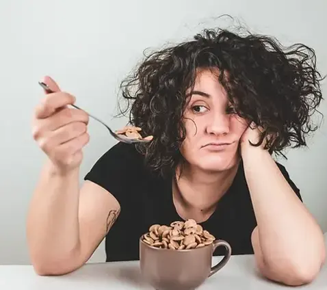 Tired-looking woman with curly hair holding a spoon and staring at a bowl of cereal with a bored expression.
