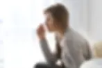 A young woman with long hair in a braid sits indoors, coughing into her fist while wearing a light gray sweater.