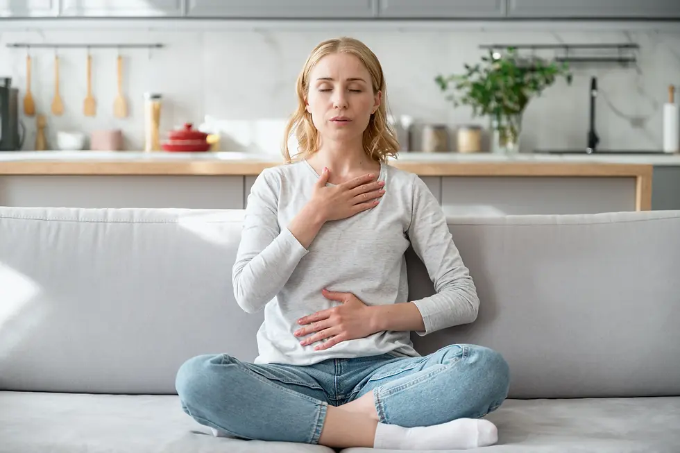 A woman sitting cross-legged on a couch with one hand on her chest and the other on her stomach, practicing deep breathing.