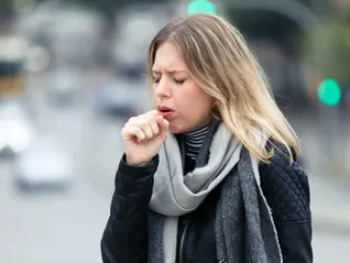 Woman outdoors coughing into fist, showing discomfort associated with chronic throat clearing while wearing scarf and jacket on city street.