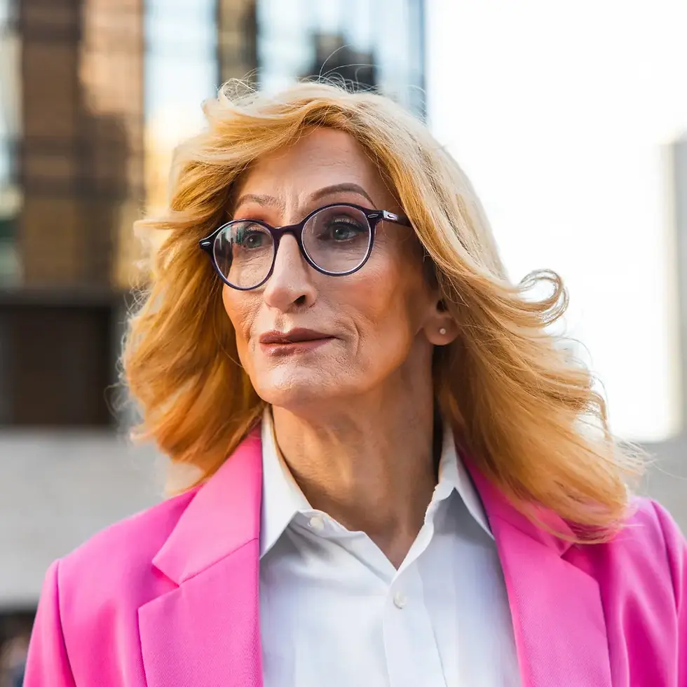 Confident transgender with blonde hair and glasses wears a bright pink blazer over a white shirt, standing outdoors in sunlight.