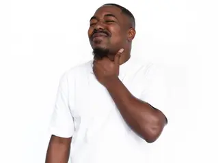 A person in a white t-shirt touches their throat with their eyes closed in discomfort showing vocal strain.